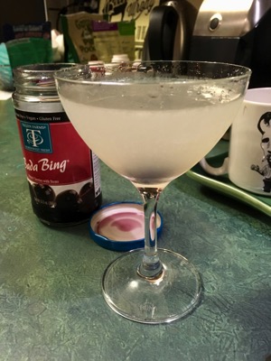 A frosty white cocktail with cherry at the bottom of the glass, a container of dark cherries behind